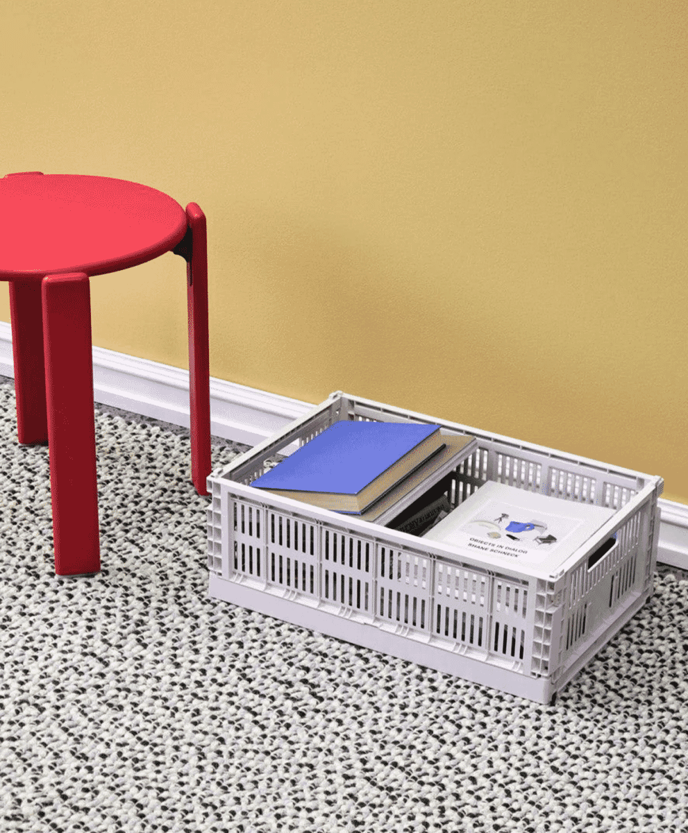 A white foldable plastic storage crate filled with books on a textured rug against a yellow wall.