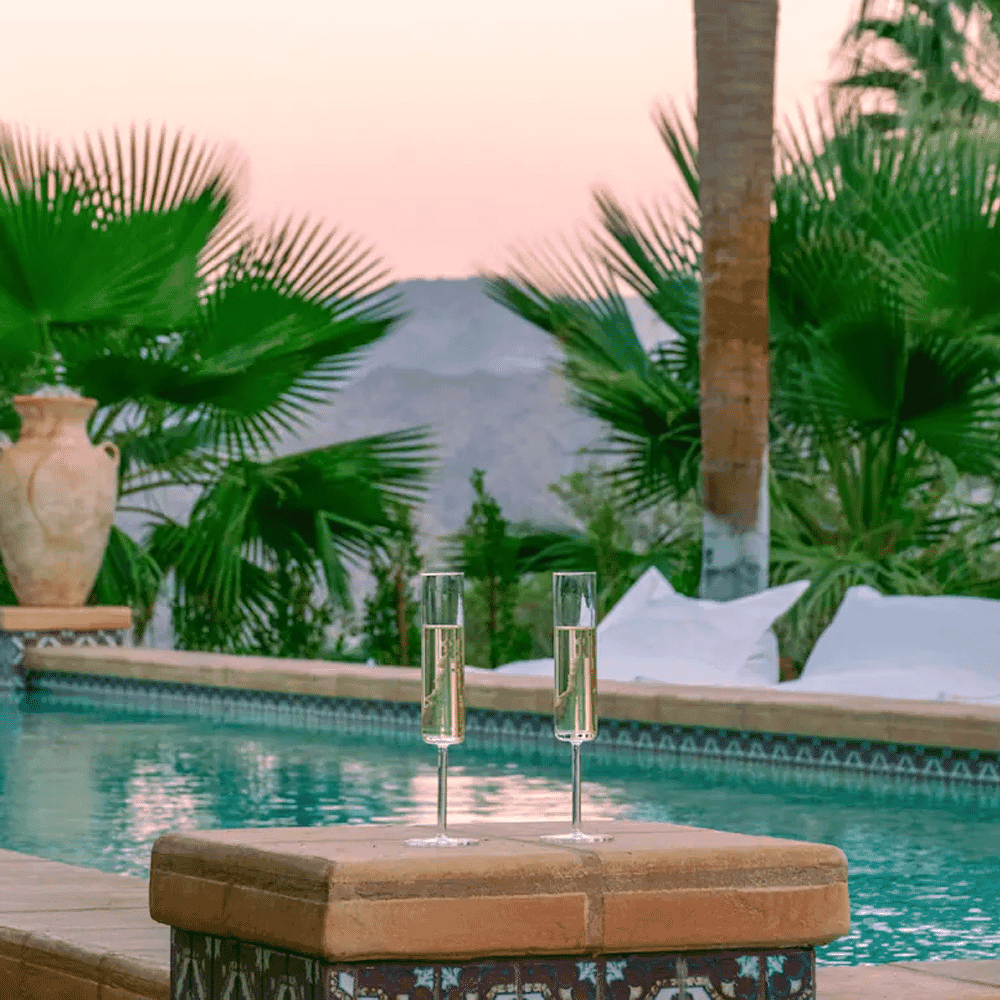 Champagne flutes on a tiled poolside ledge with mountains and palm trees in the distance.