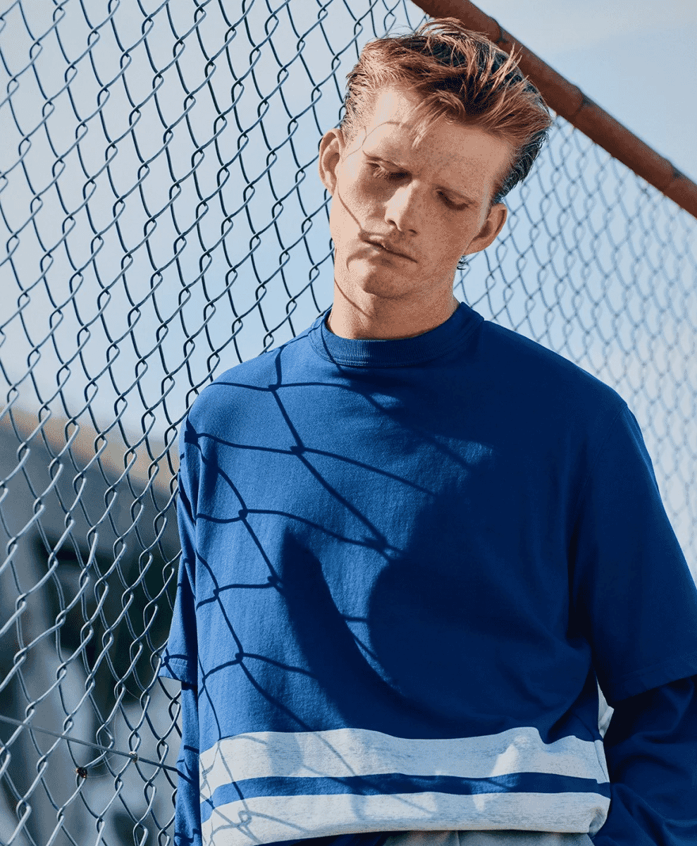 A man in a blue long-sleeve crewneck with white horizontal stripes against a chain-link fence.