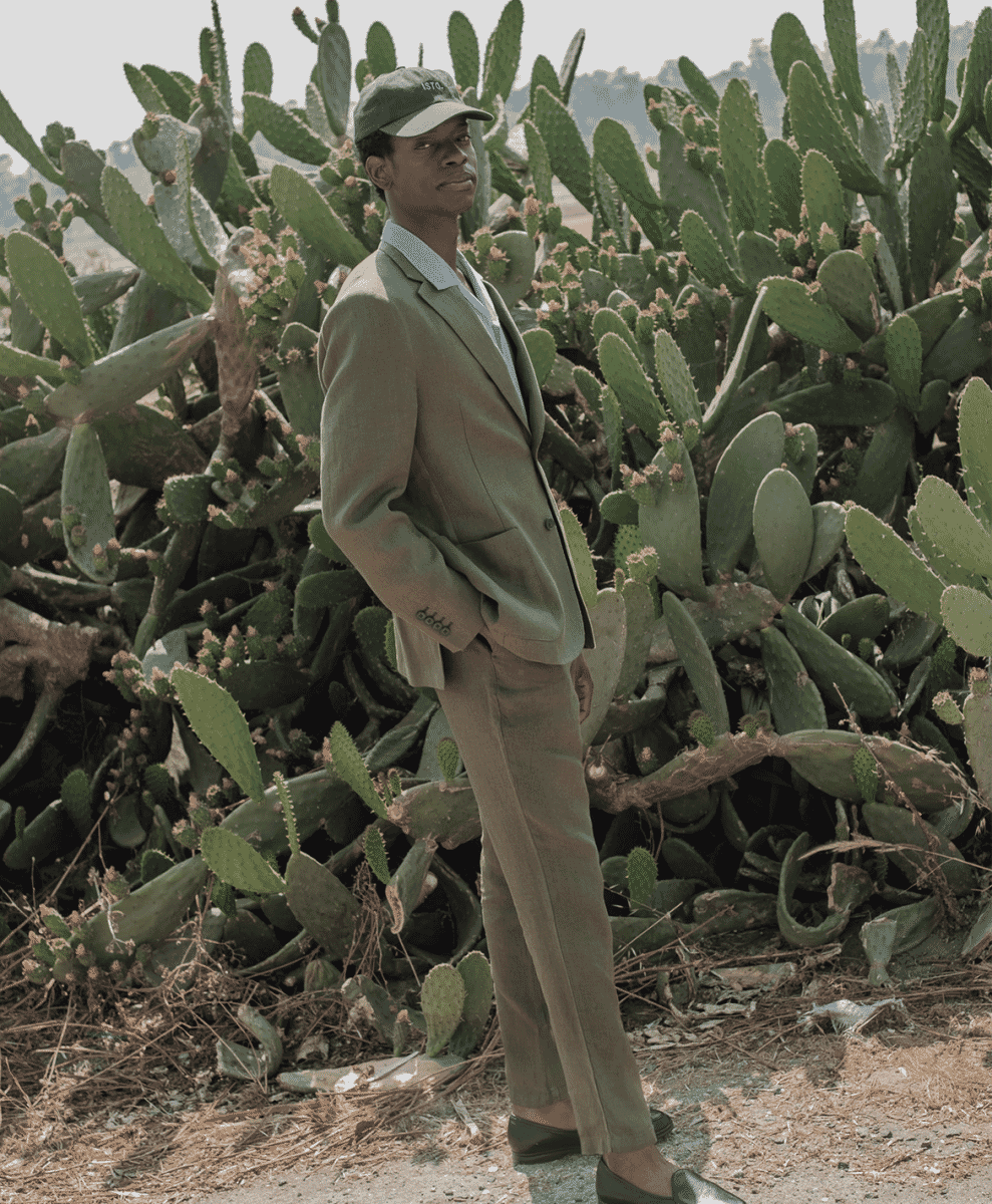 A man in a muted olive-green suit and matching cap poses against a backdrop of cacti.