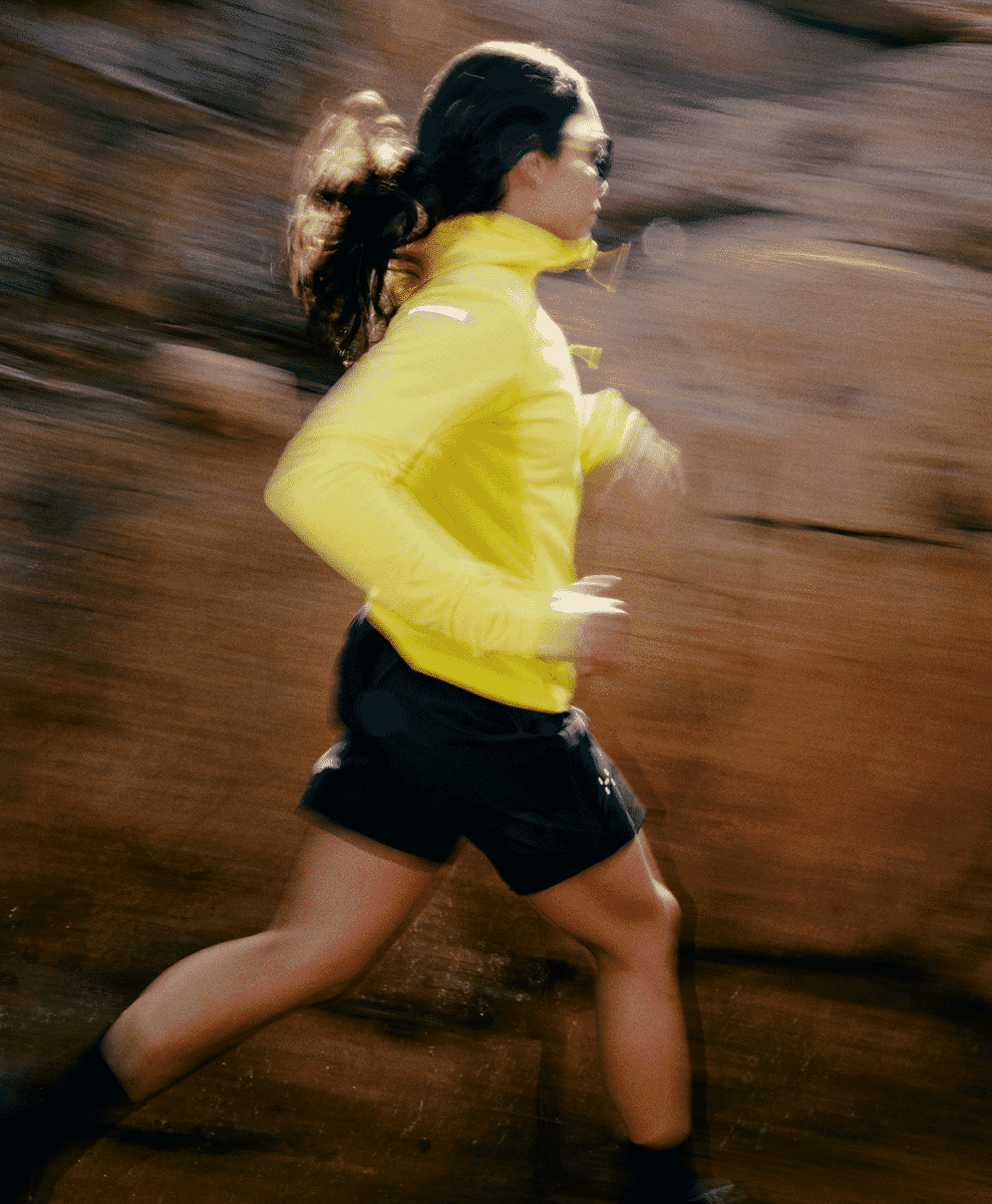 A woman in a bright yellow athletic jacket running through a rocky outdoor environment with motion blur.