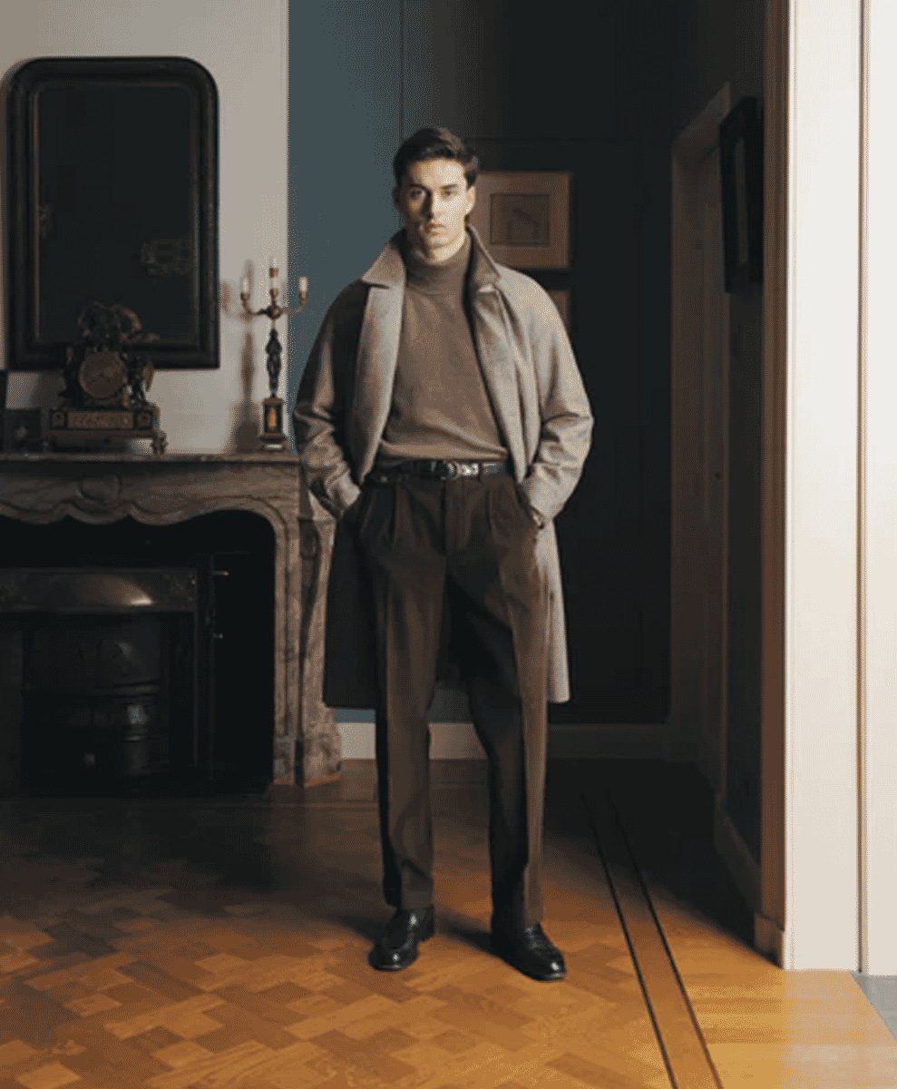 A man in a beige coat, brown turtleneck, and pleated trousers in a classic room.