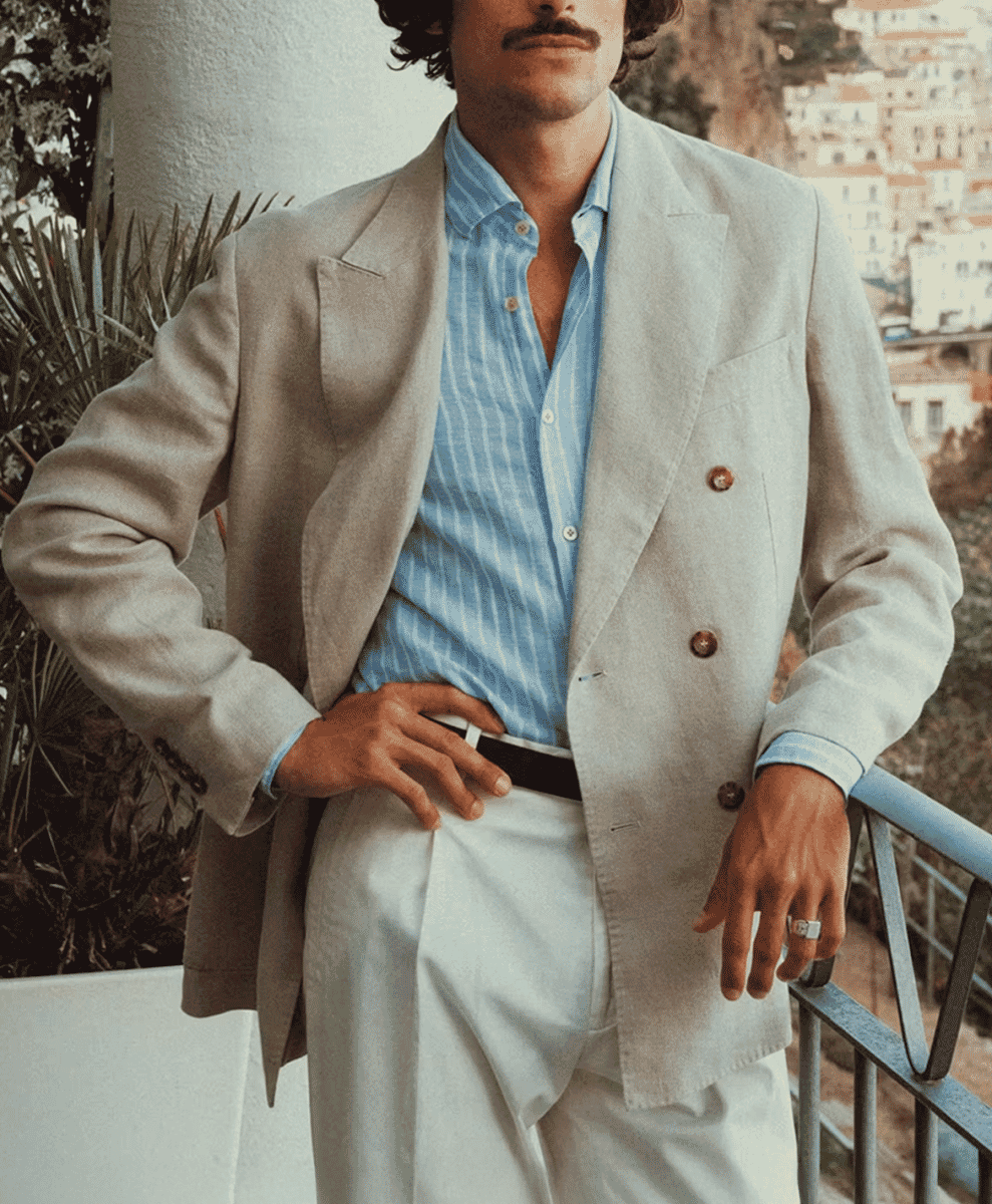 Man in a light beige double-breasted blazer and white trousers on a balcony overlooking a Mediterranean coastal town.