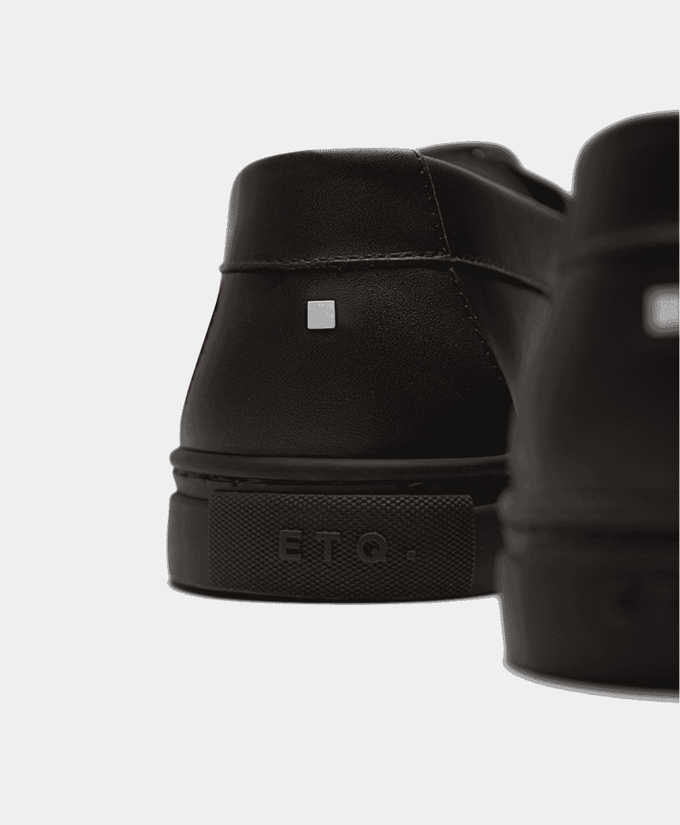 Close-up of the metallic square logo on the heel of a black leather shoe.