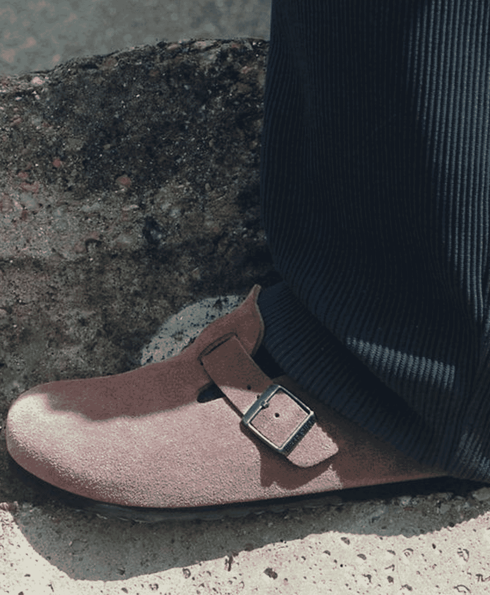 Close-up of a person wearing dusty rose suede clogs and dark ribbed corduroy pants on a concrete surface.