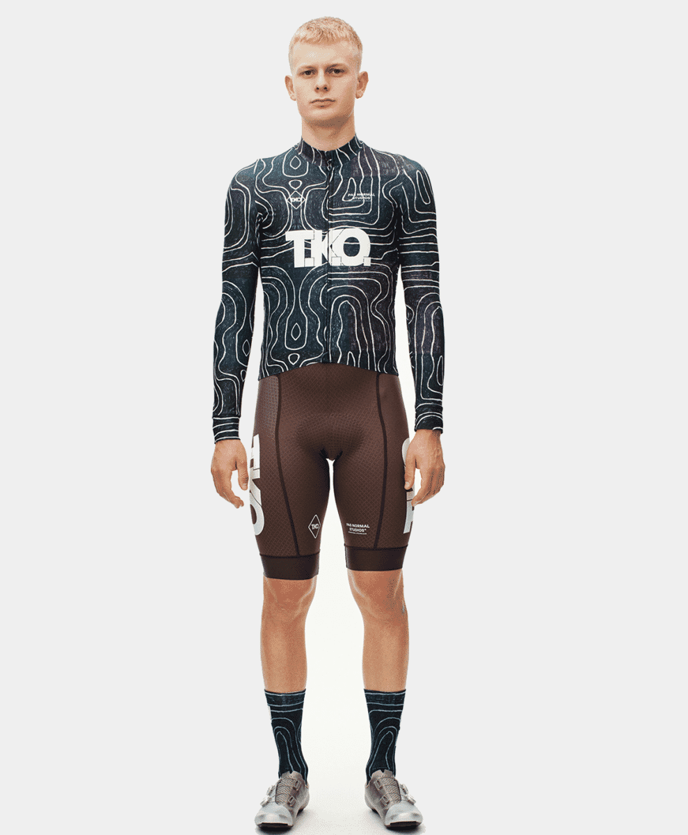 Male model wearing a topographic print long-sleeve jersey and brown bib shorts against a white background.
