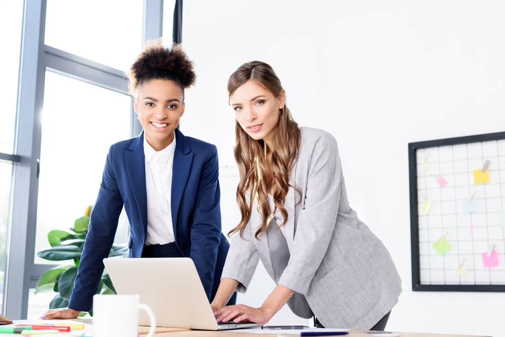 Two women in navy and gray blazers collaborating at a desk with a laptop.