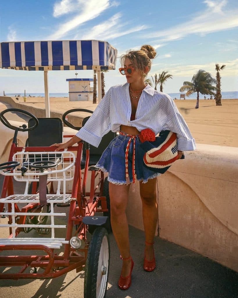 Woman in a striped shirt and red heels posing by a bicycle at the beach.