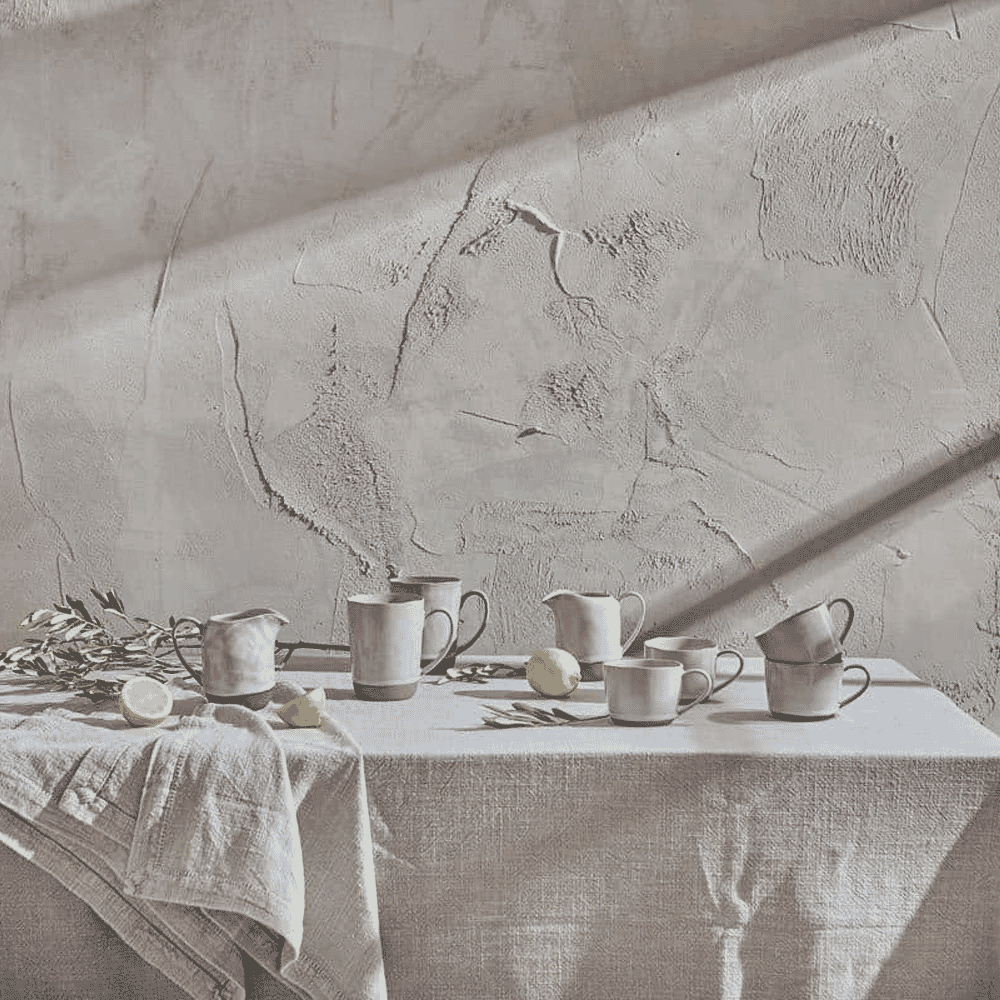A still life of rustic ceramic mugs and pitchers in natural light on a linen cloth.