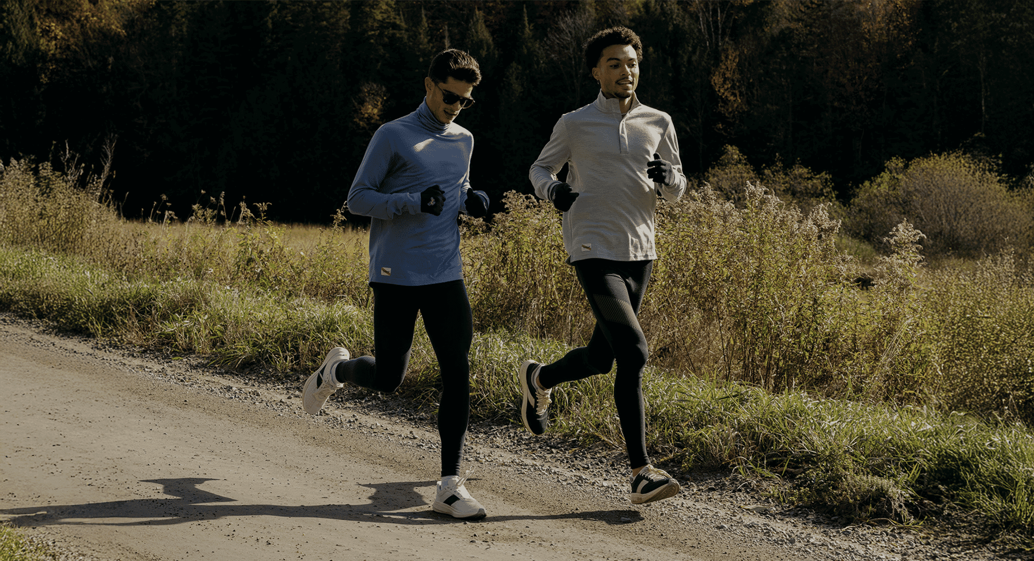 Two men running on a sunlit dirt path through a forest with autumn foliage.