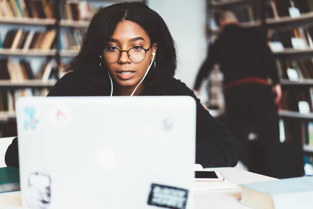 A woman with glasses and earphones at a library desk, embodying a casual academic style.