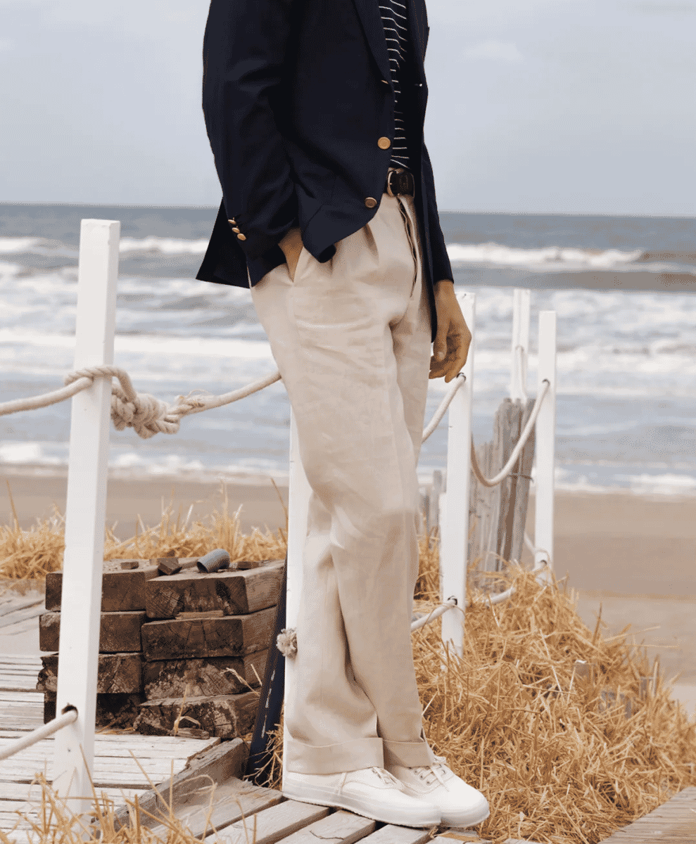 A man in beige single-pleat linen trousers and white sneakers on a coastal boardwalk