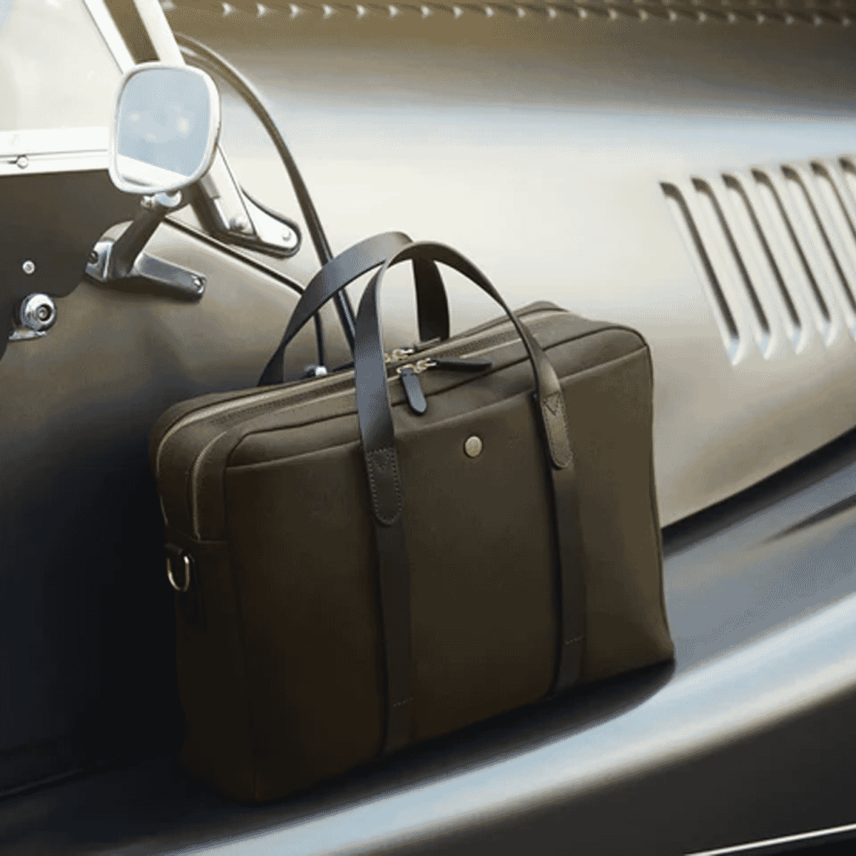 A dark brown leather briefcase resting on the silver hood of a classic vintage car.