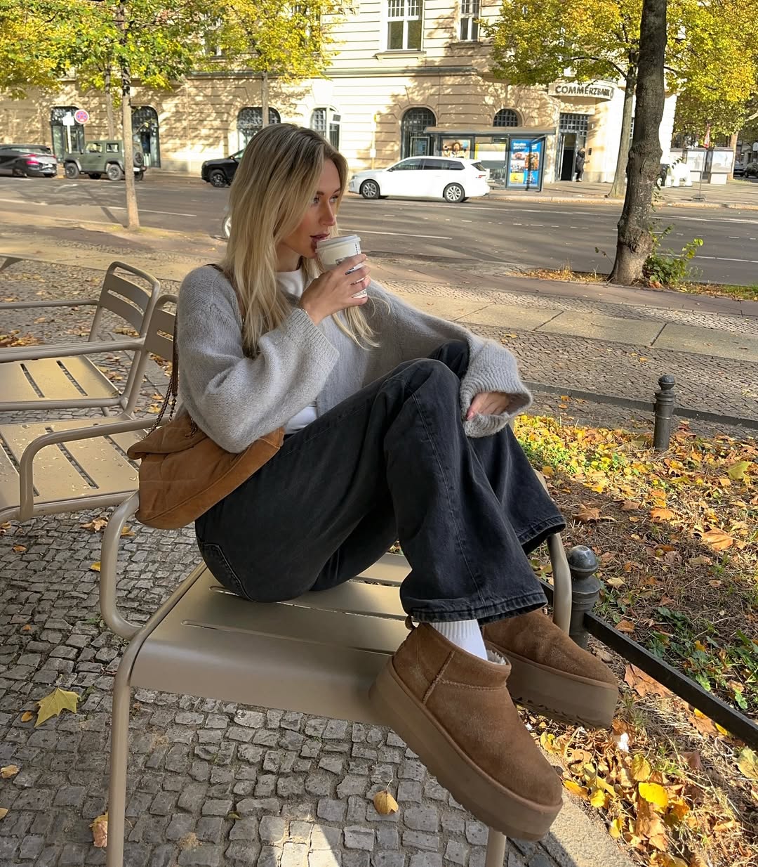 Blonde woman in a grey knit sweater and dark wide-leg jeans wearing platform boots while sitting on a city bench.