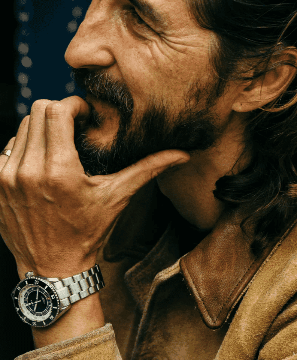 A man in a brown suede jacket wearing a silver-toned watch in a contemplative pose.