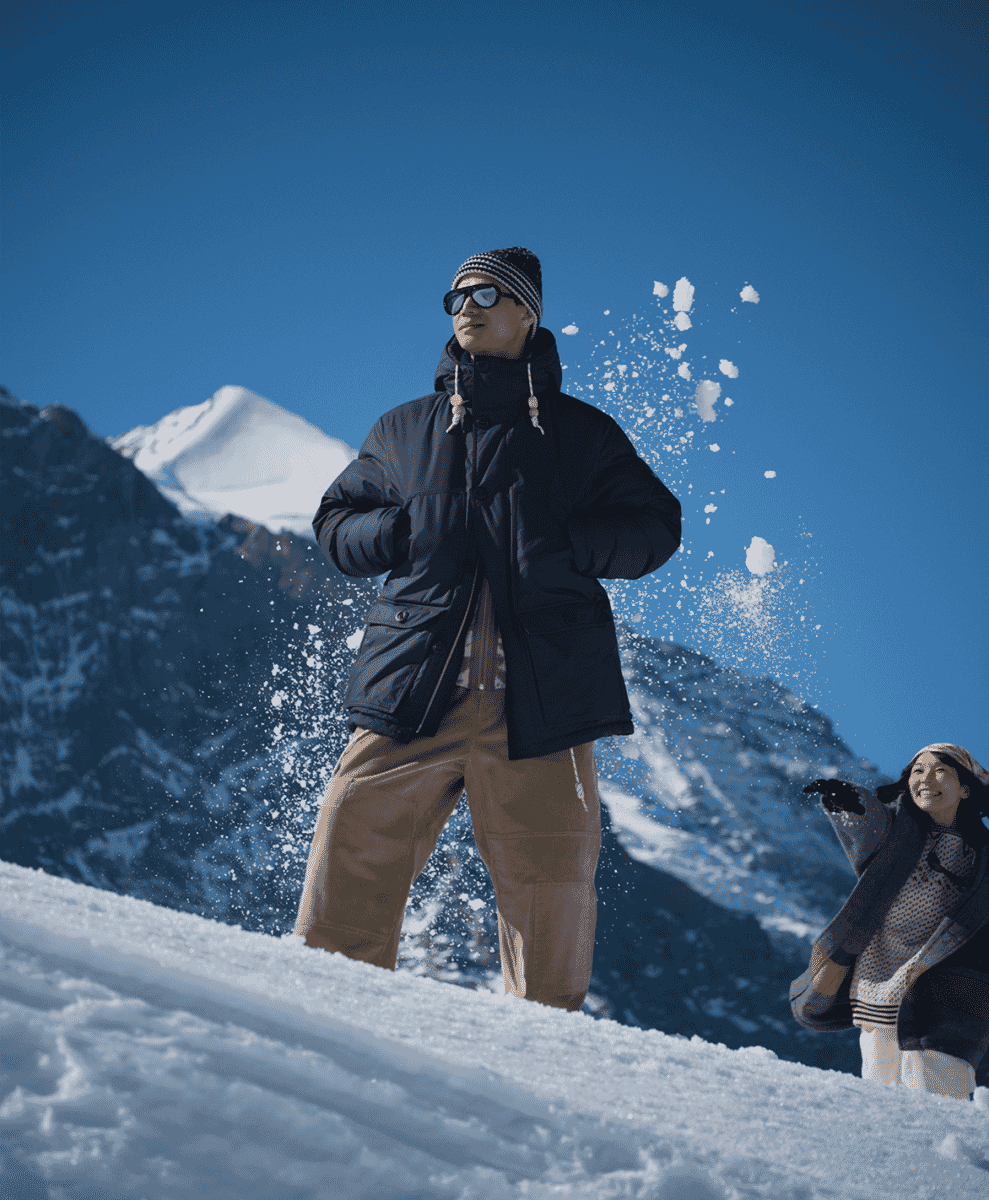 Man in a black puffer jacket, striped beanie, and sunglasses standing on a snowy mountain slope.