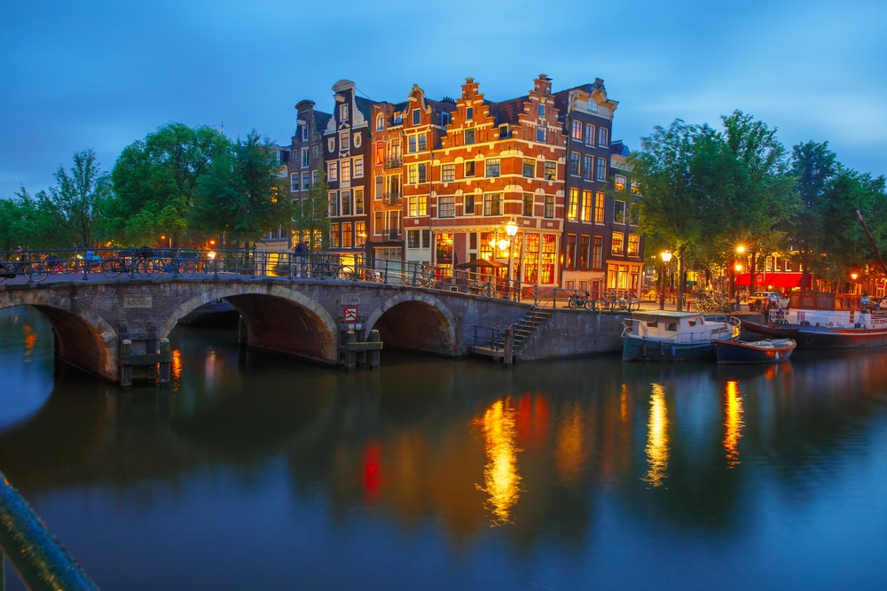Evening view of an Amsterdam canal with historic gabled houses and a bridge reflected in the water.
