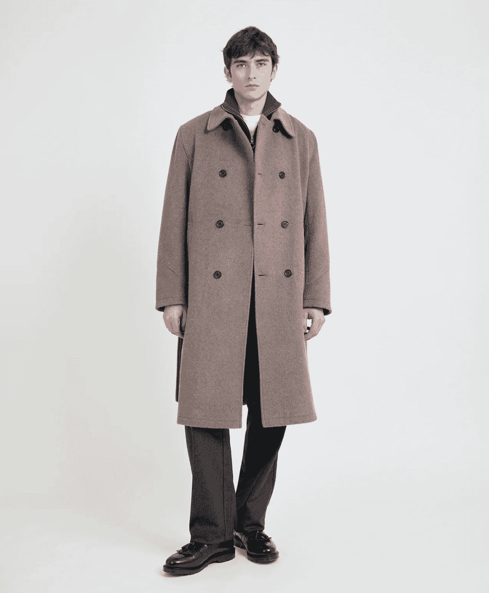 A man wearing a long, double-breasted brown wool coat in a full-body studio shot.