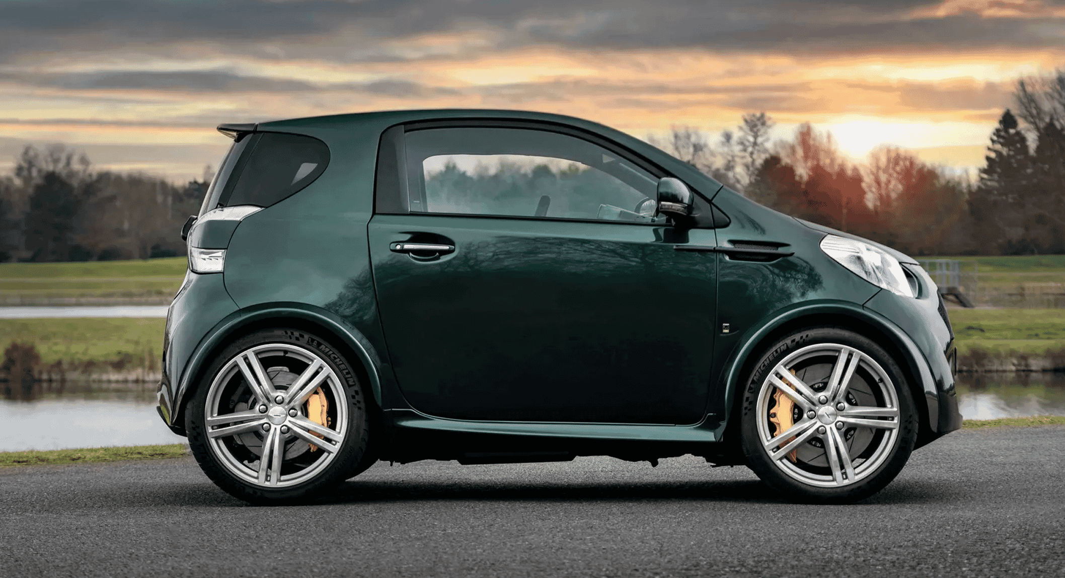 Sleek dark green compact car with yellow brake calipers parked near a pond at sunset.