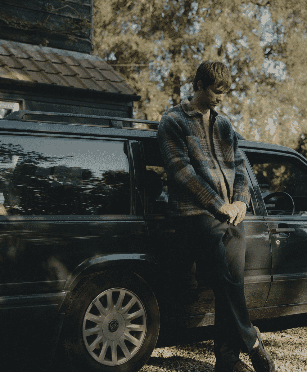 A man in a plaid wool coat leaning against a vintage Volvo station wagon outdoors.