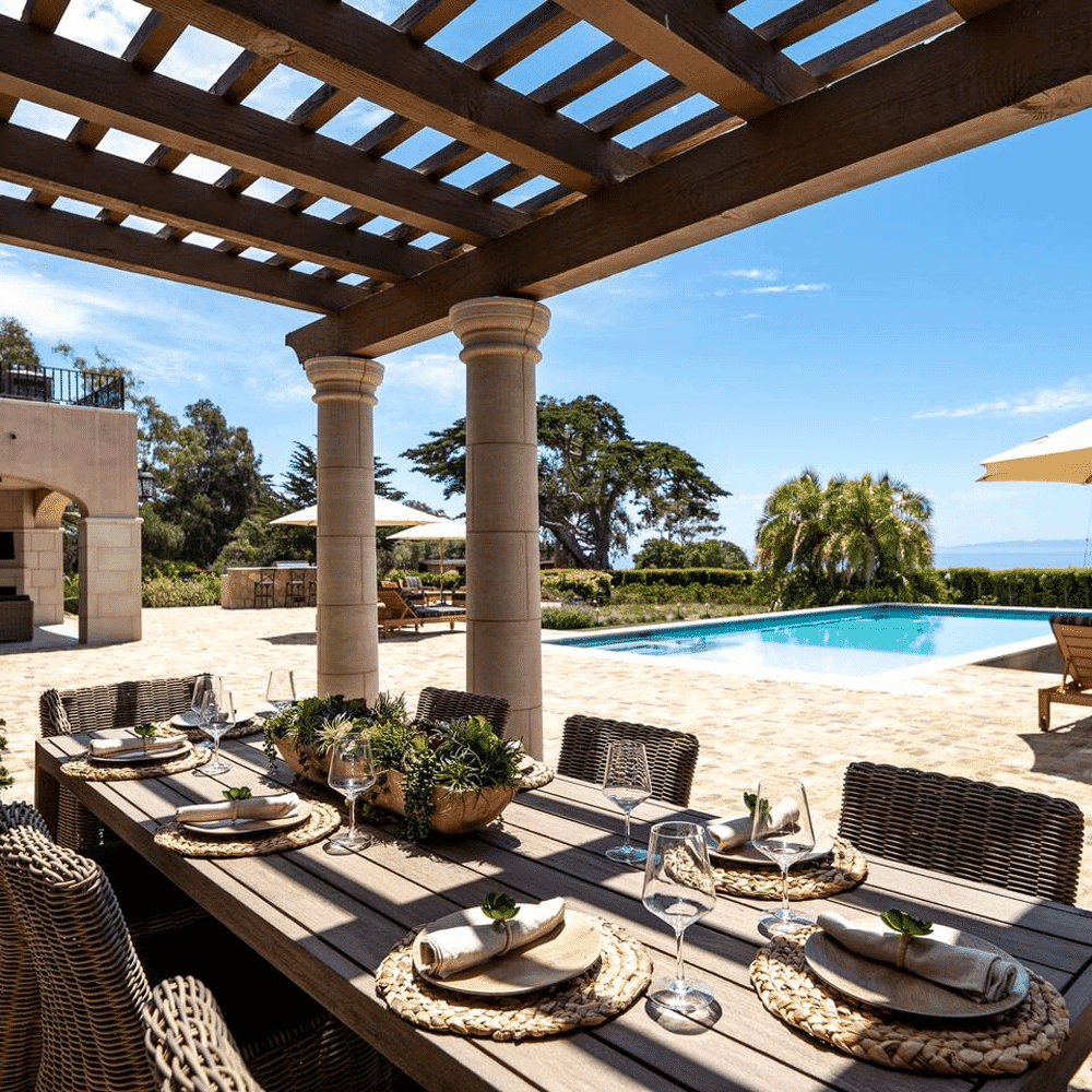 An outdoor patio dining setup under a wooden pergola overlooking a pool and the ocean.