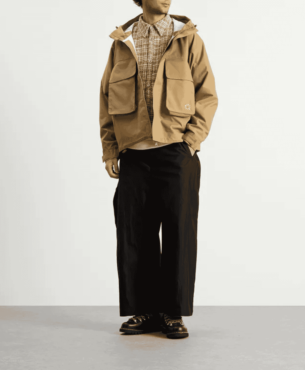 Model in a tan hooded utility jacket, patterned shirt, and black wide-leg trousers.