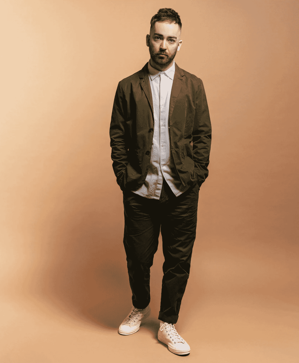 A man in a brown textured blazer, dark trousers, and white sneakers.