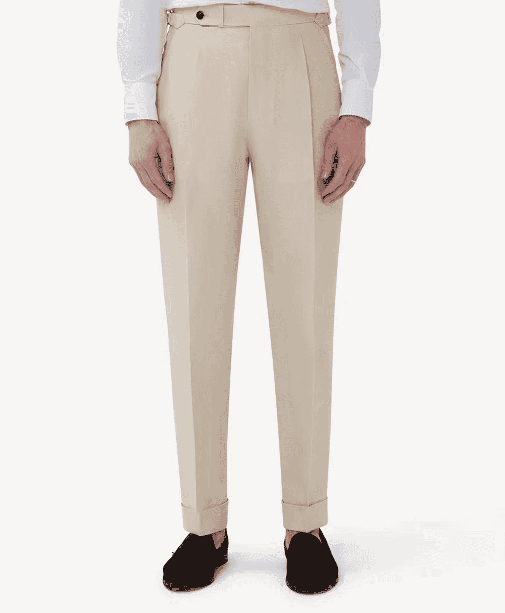 Man modeling beige pleated trousers with cuffed hems and dark loafers against a white studio background.