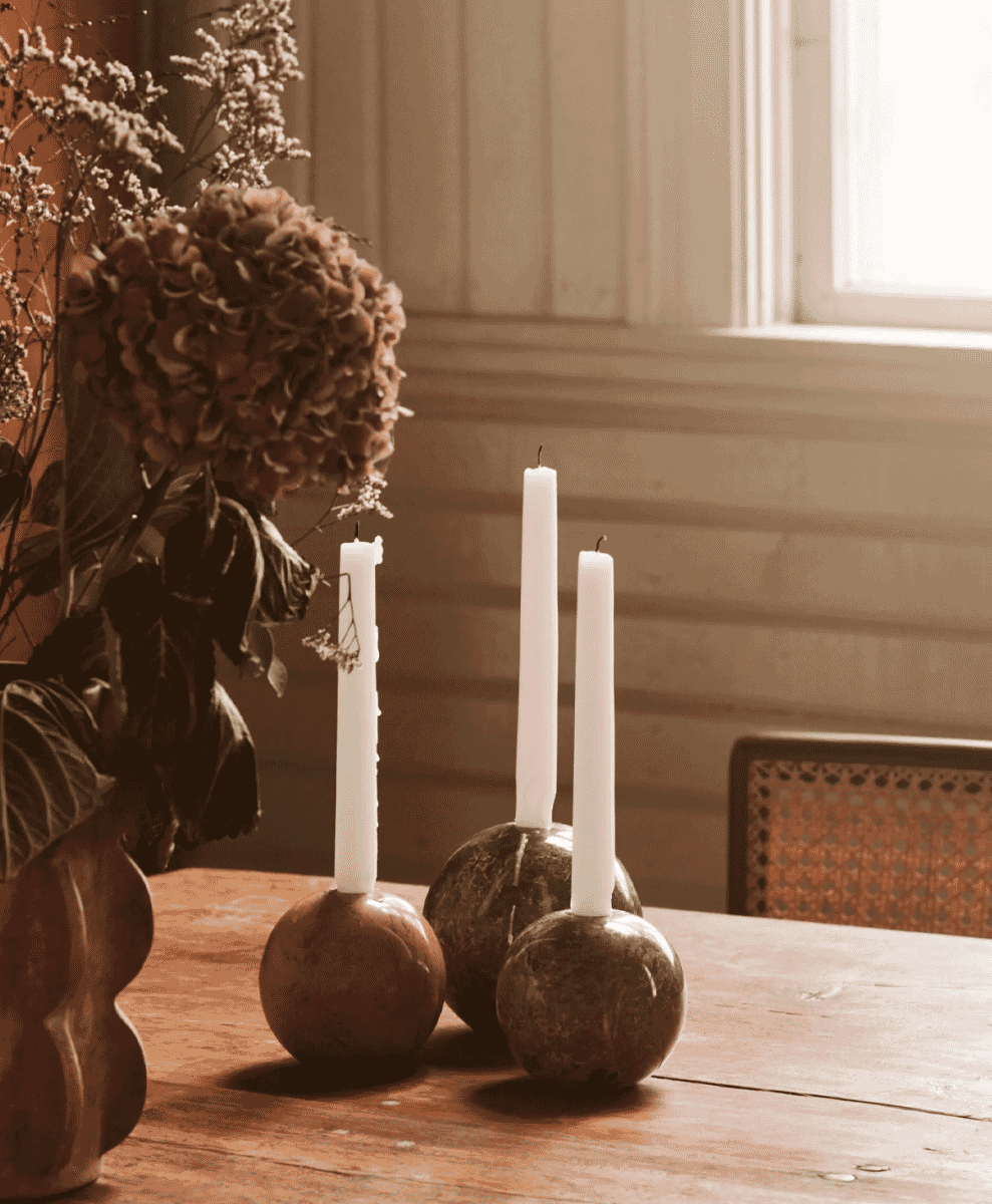 Three spherical stone candle holders with white candles on a wooden table beside dried flowers.