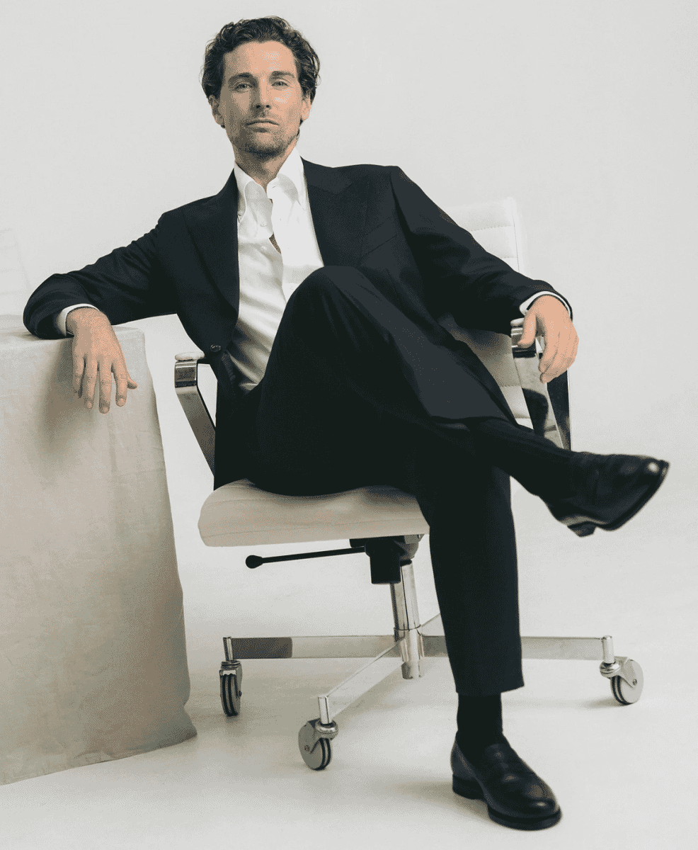 A man in a sharp black suit sitting confidently on a modern office chair.