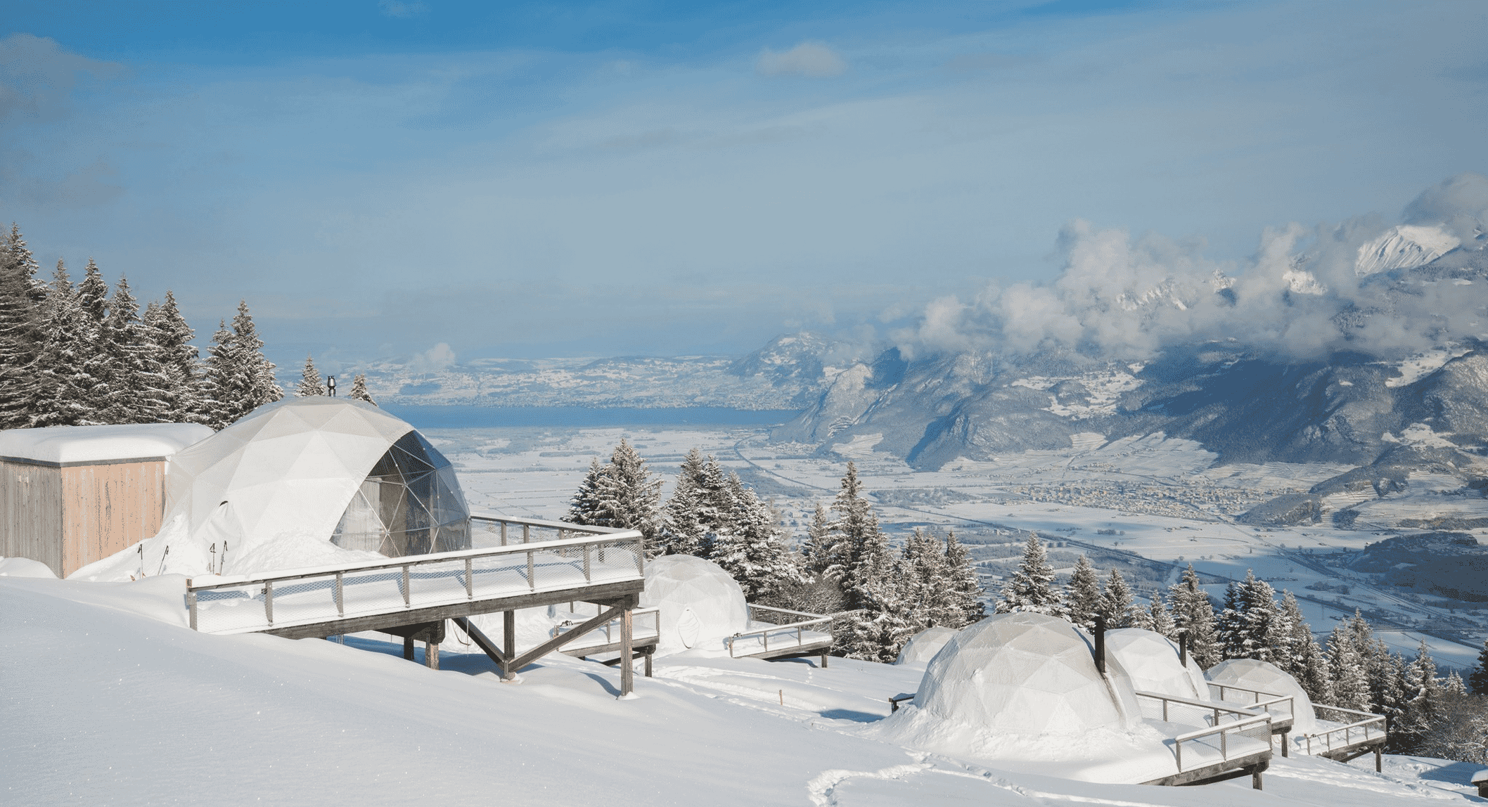 Snow-covered geodesic domes on a mountain slope surrounded by evergreen trees.