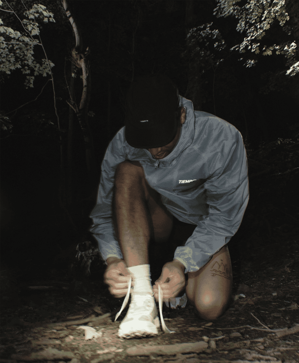 A man in a reflective Tiempos jacket crouching in a dark forest at night.