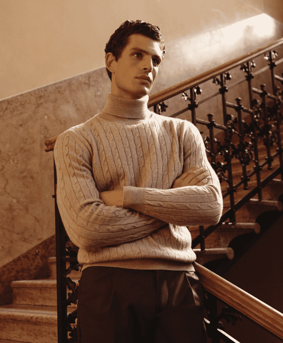 A man wearing a heavy cream-colored cable-knit turtleneck standing on an ornate indoor staircase.