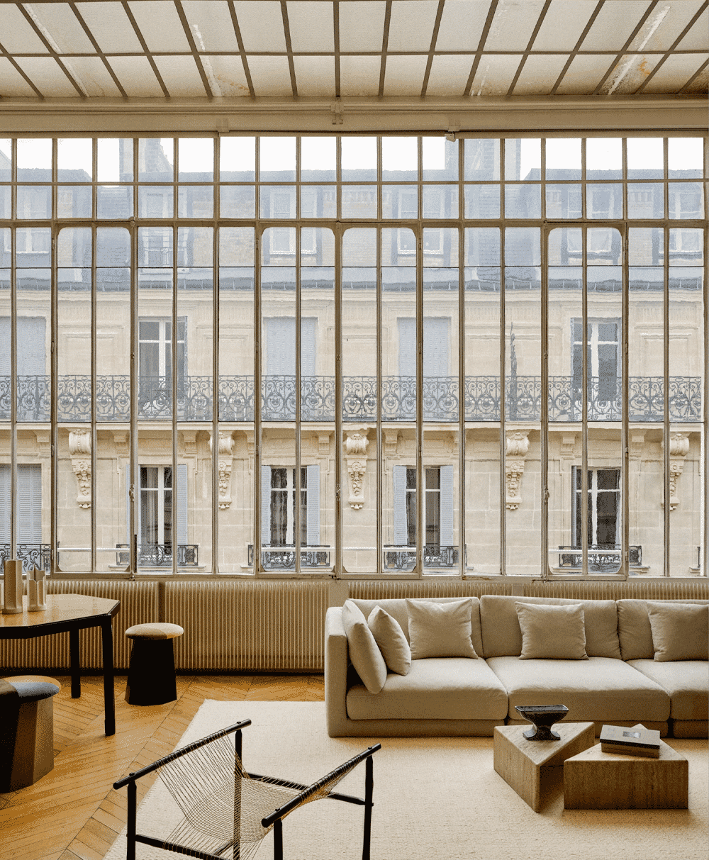 A minimalist living room with a beige sofa and large windows overlooking Parisian buildings.