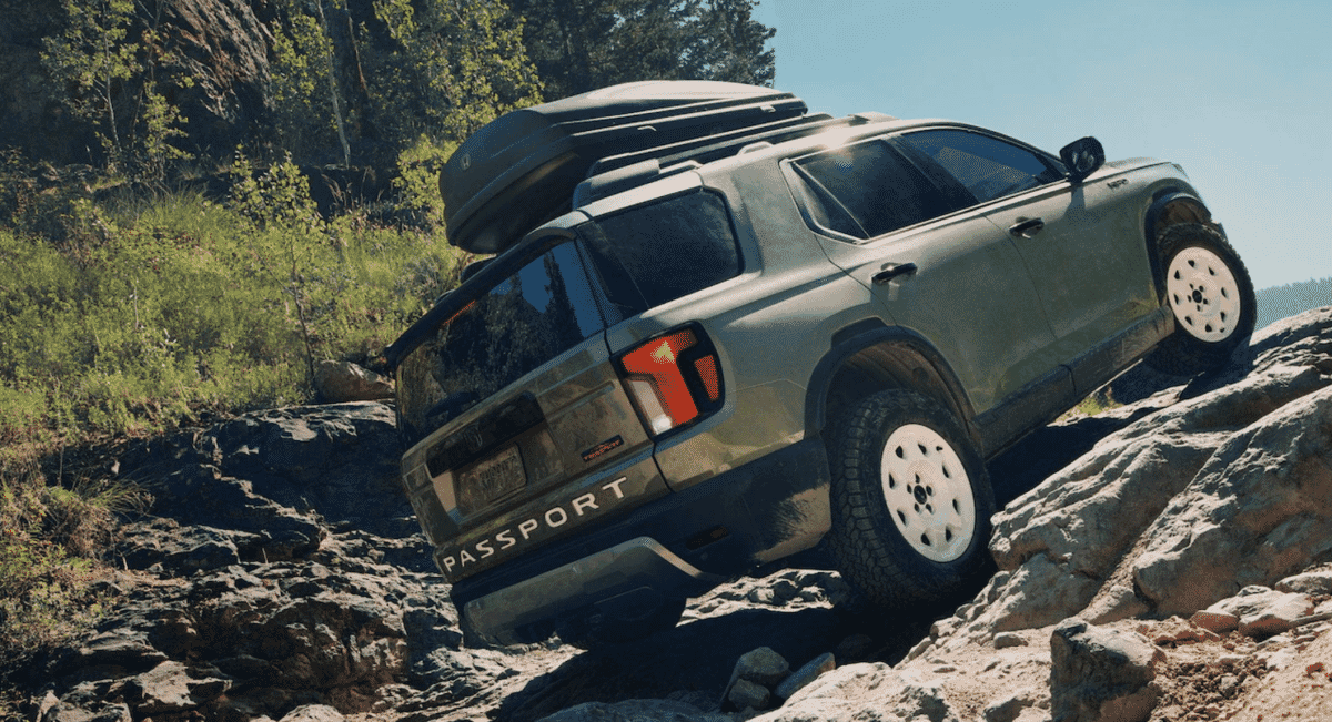 A silver SUV with a roof cargo box navigating a forested mountain trail.
