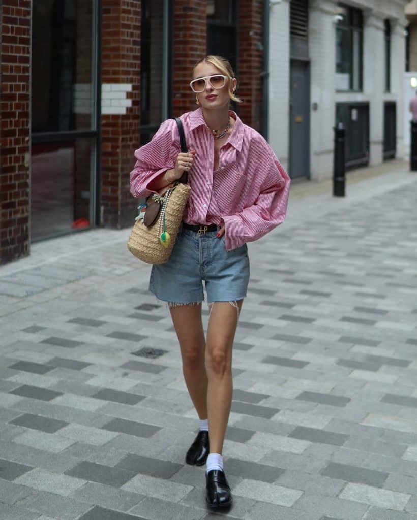 Woman in an oversized pink striped shirt and denim shorts walking on a brick street.