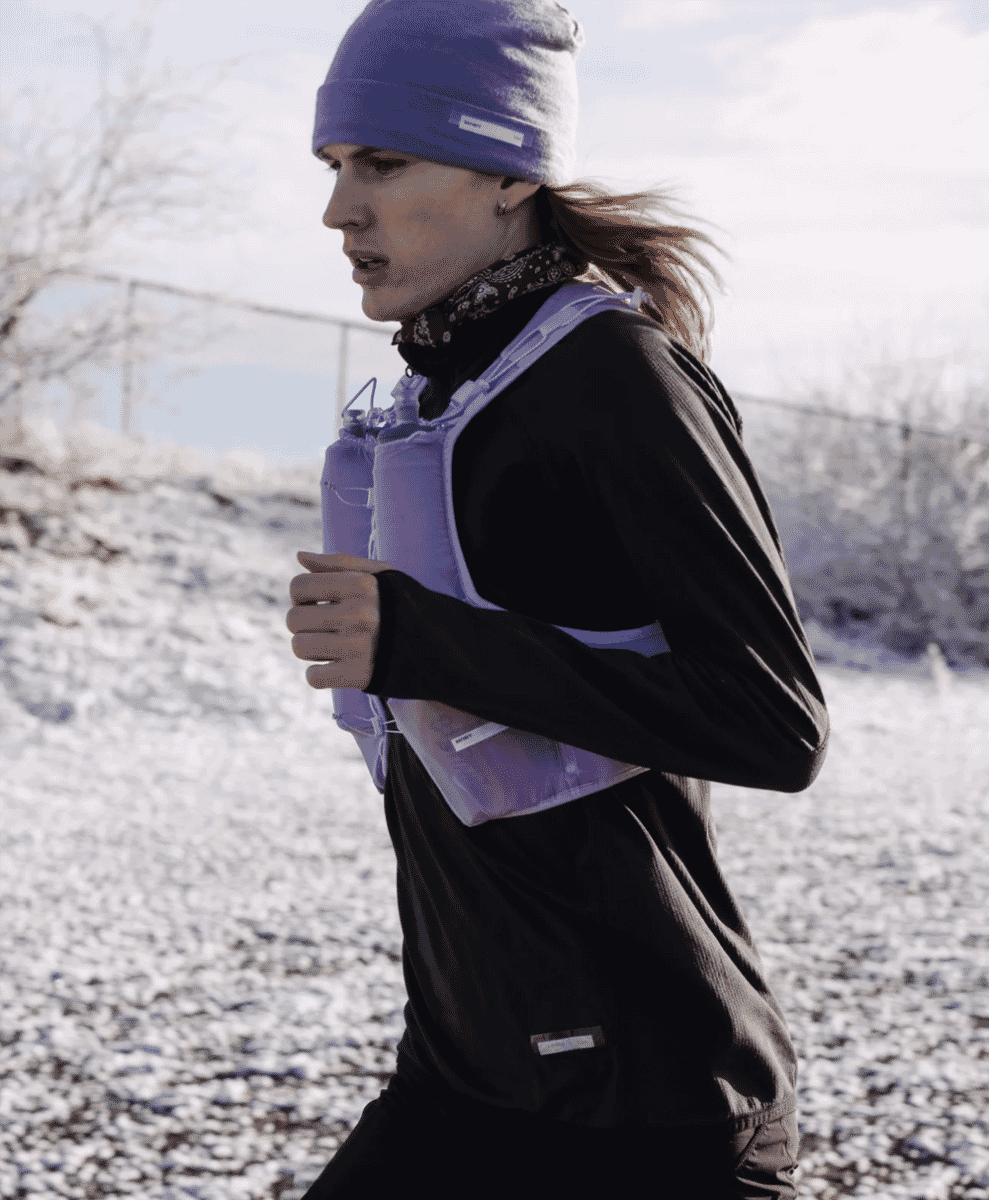 A woman runs in snow wearing a purple hydration vest and cold-weather running gear.