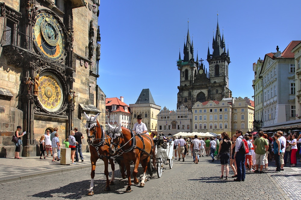 Tourists walking through a historic European square with horse-drawn carriages and Gothic architecture.