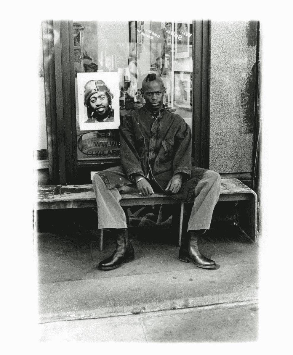 A man with a mohawk in a loose denim jacket sitting in an urban storefront setting.