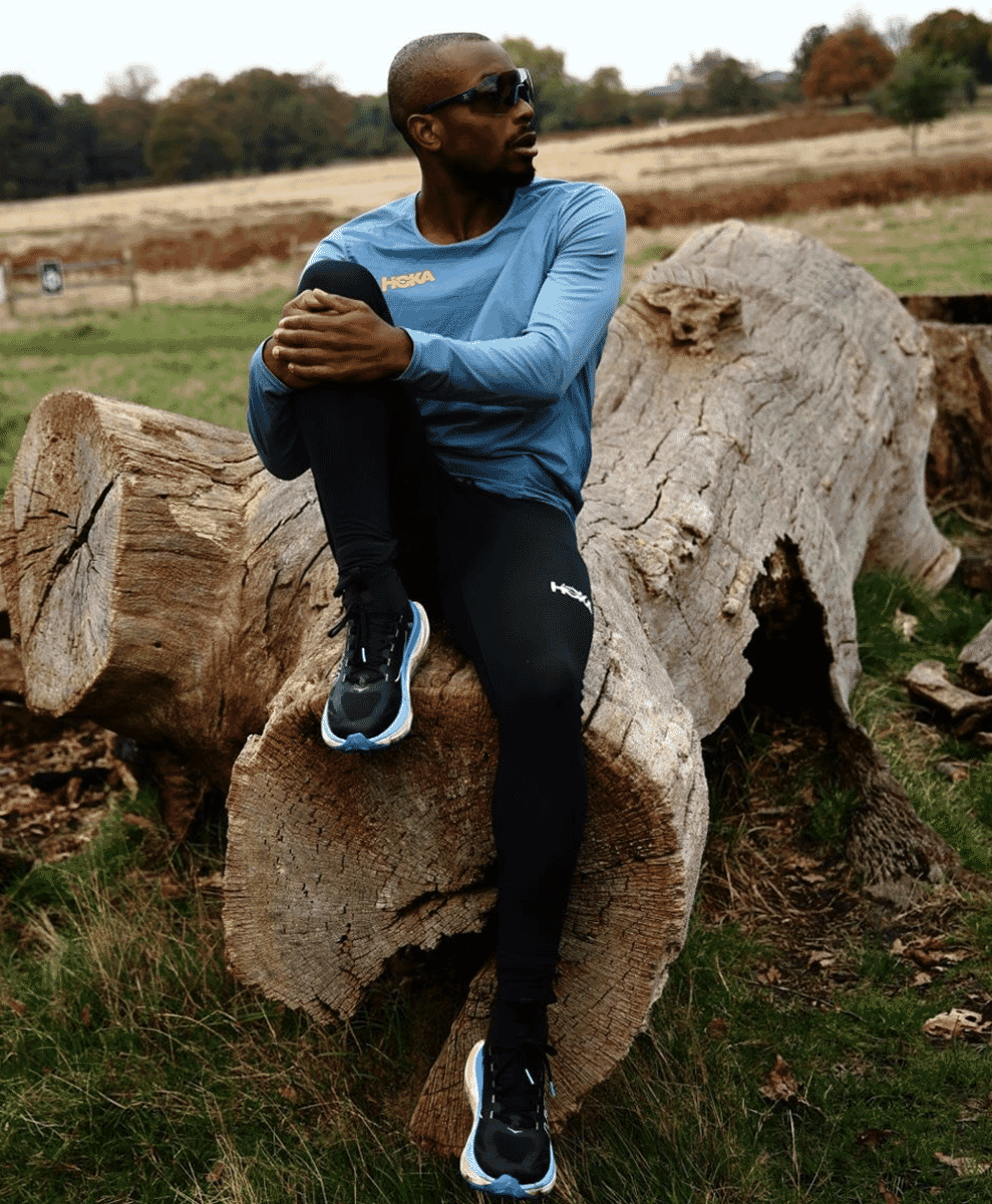 A runner resting on a log in a blue HOKA long-sleeve and black compression leggings in an autumn landscape.