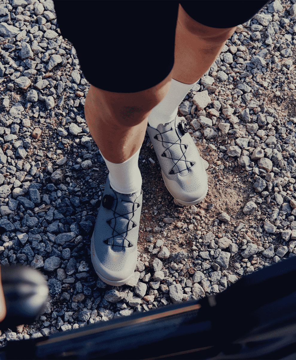 Cyclist standing on a gravel path wearing light grey QUOC cycling shoes and white socks.