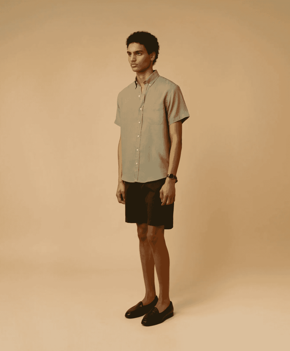 Full body shot of a man in an olive-beige short-sleeve button-up with dark shorts and black loafers.
