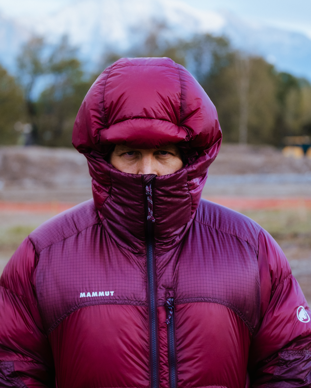 A man in a maroon Mammut puffer jacket zipped high against a cloudy sky.