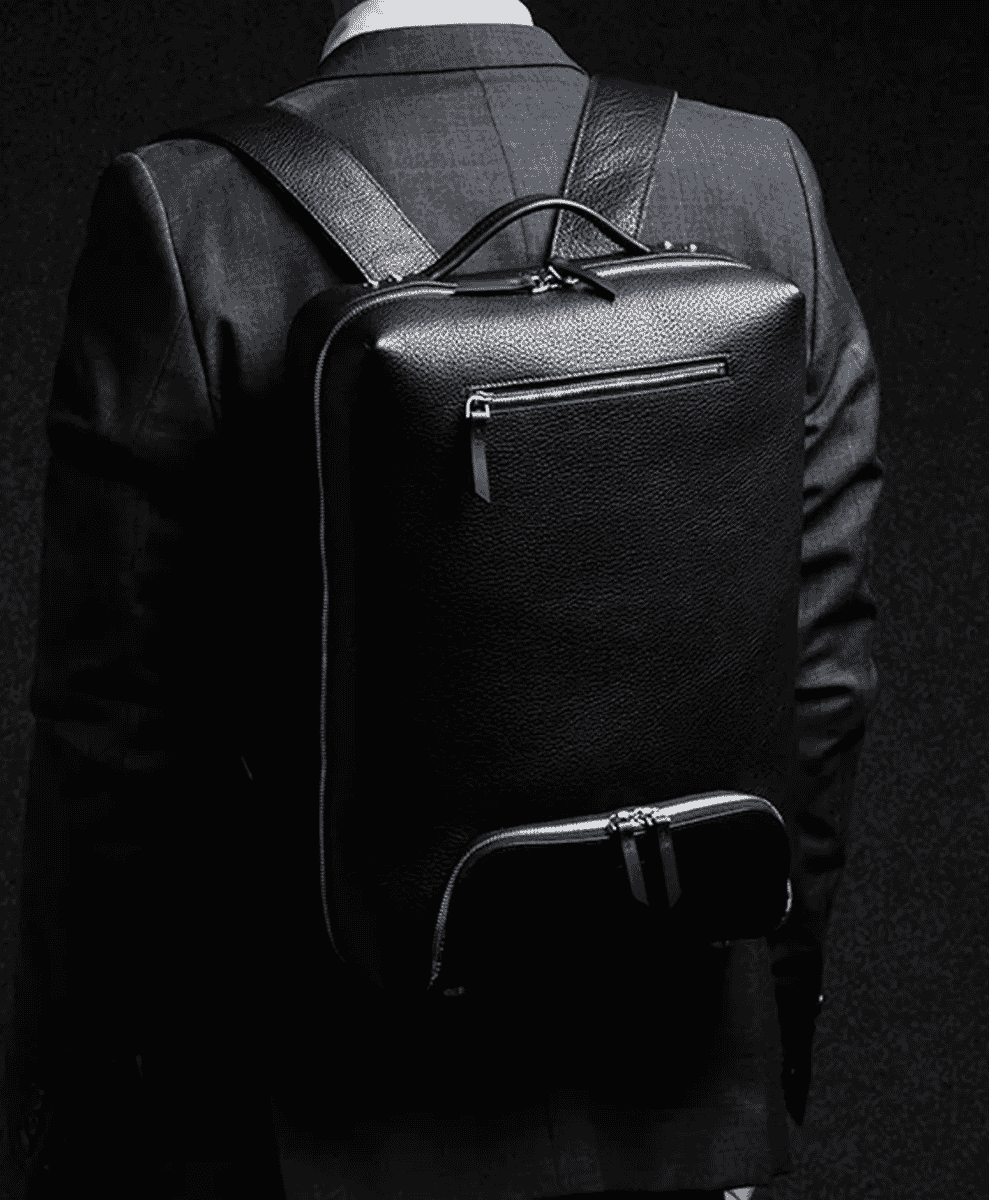 A man in a dark tailored suit wearing a textured black leather backpack.