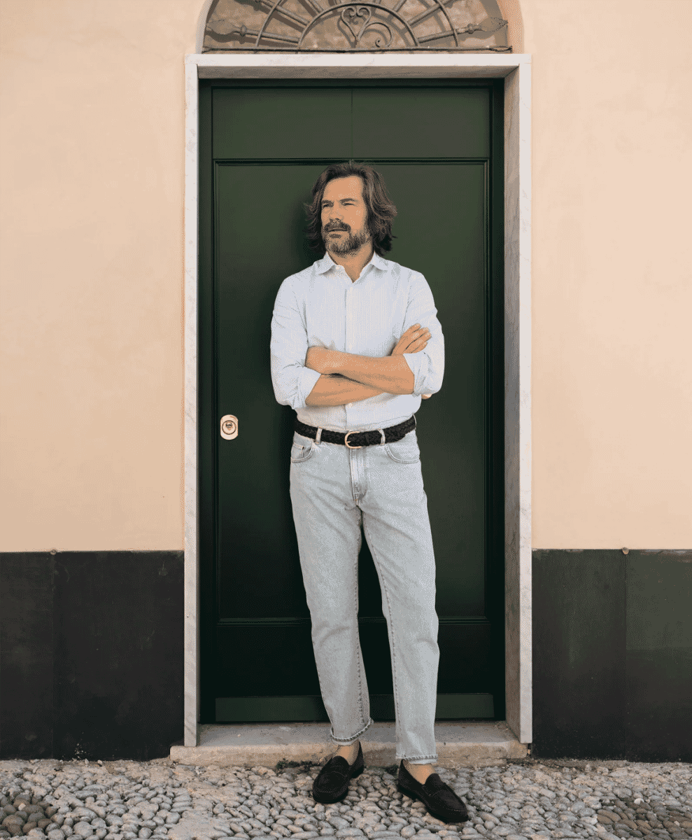 A man in a light blue striped Oxford shirt and light-wash denim jeans standing in front of a green door.