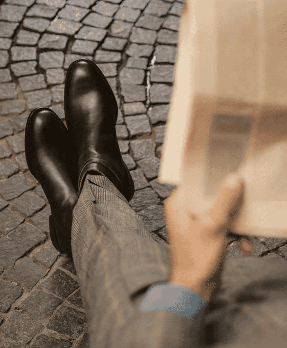 A man in a grey plaid suit and black leather boots sitting on a cobblestone street reading a newspaper.