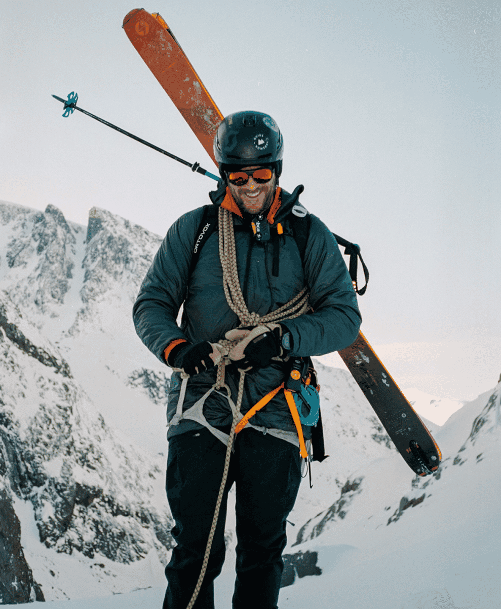 A smiling skier in a helmet and goggles with skis over his shoulder on a snowy mountain ridge.