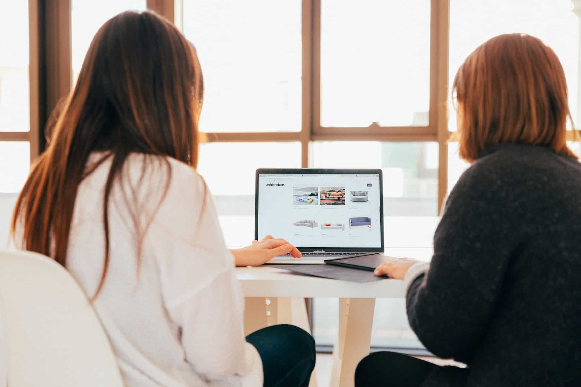 Two women in casual knitwear working together on a laptop in a collaborative workspace.