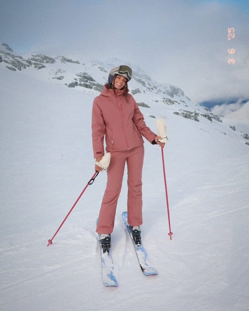 Woman in a dusty rose pink ski suit and white gloves on a snowy slope.