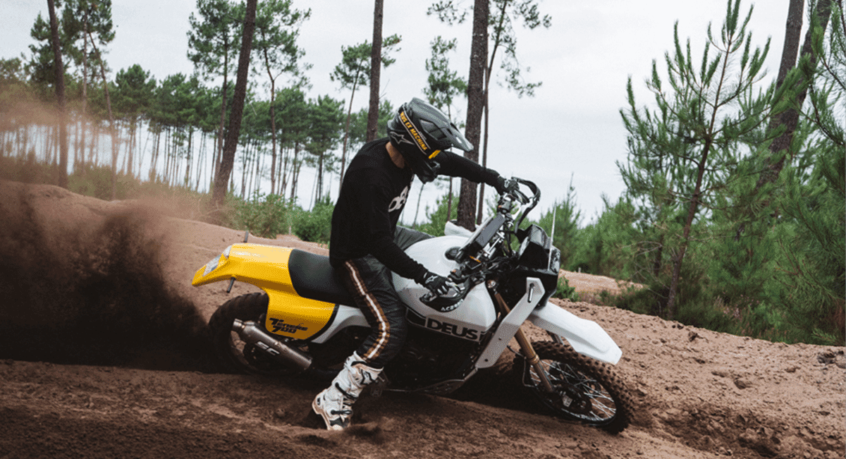 A rider in a black Deus jersey and striped motocross pants leaning into a turn in a forest.